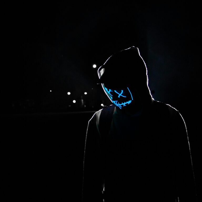 Silhouette of a person in a hoodie wearing a glowing LED mask against a dark background.