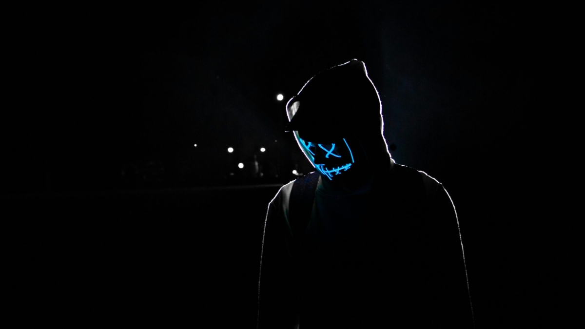 Silhouette of a person in a hoodie wearing a glowing LED mask against a dark background.
